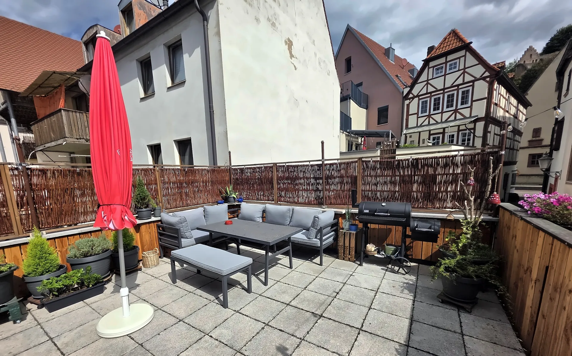 Terrasse mit grauen Loungesofas, Tisch, Grill und rotem Sonnenschirm, dahinter Fachwerkhäuser